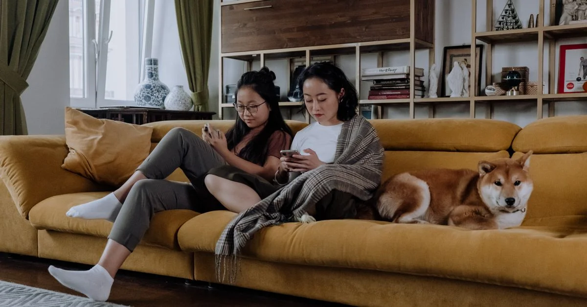 Deux femmes assises sur un canapé avec un chien Shiba Inu, utilisant des smartphones dans un salon confortable.