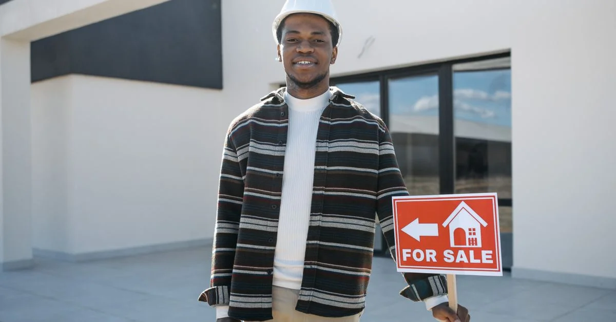 Jeune homme noir agent immobilier tenant un panneau À VENDRE devant une maison moderne, souriant.