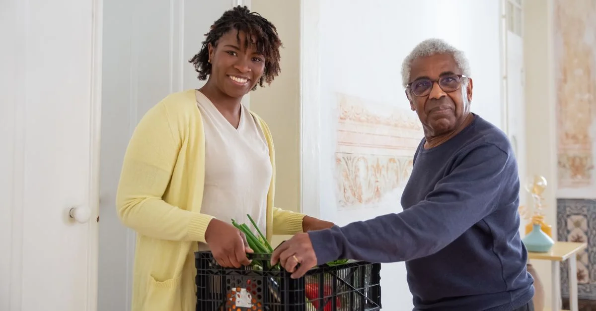 Une femme souriante aide un homme âgé avec une livraison de nourriture à l'intérieur, promouvant le soutien communautaire et le soin.