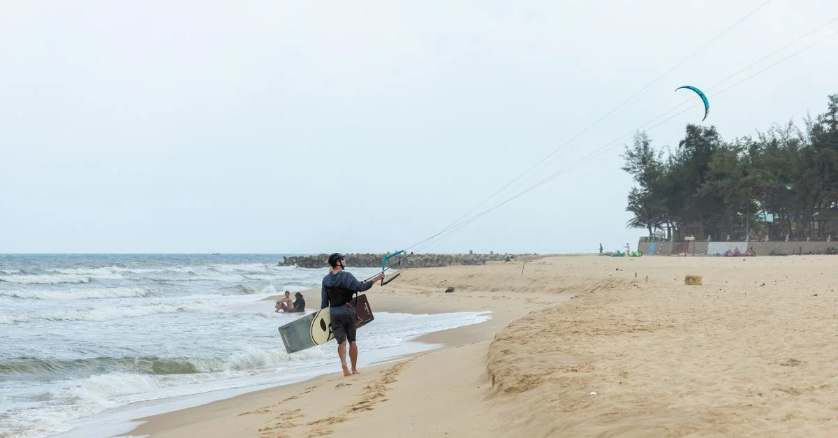 Un homme marchant sur une plage de sable portant un kiteboard, avec un cerf-volant volant au-dessus dans un cadre côtier.