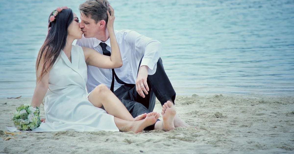 Couple en tenue de mariage partageant un bisou romantique sur une plage de sable.