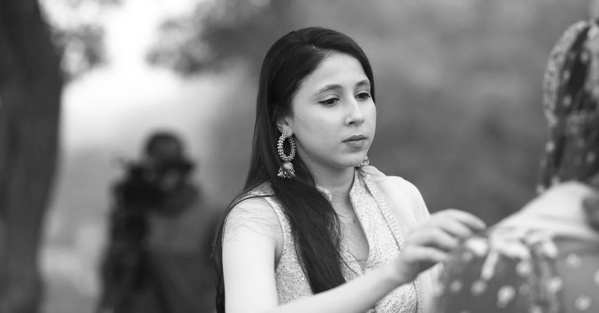 Une femme élégante ajuste une robe en plein air tandis qu'un cameraman filme en niveaux de gris.