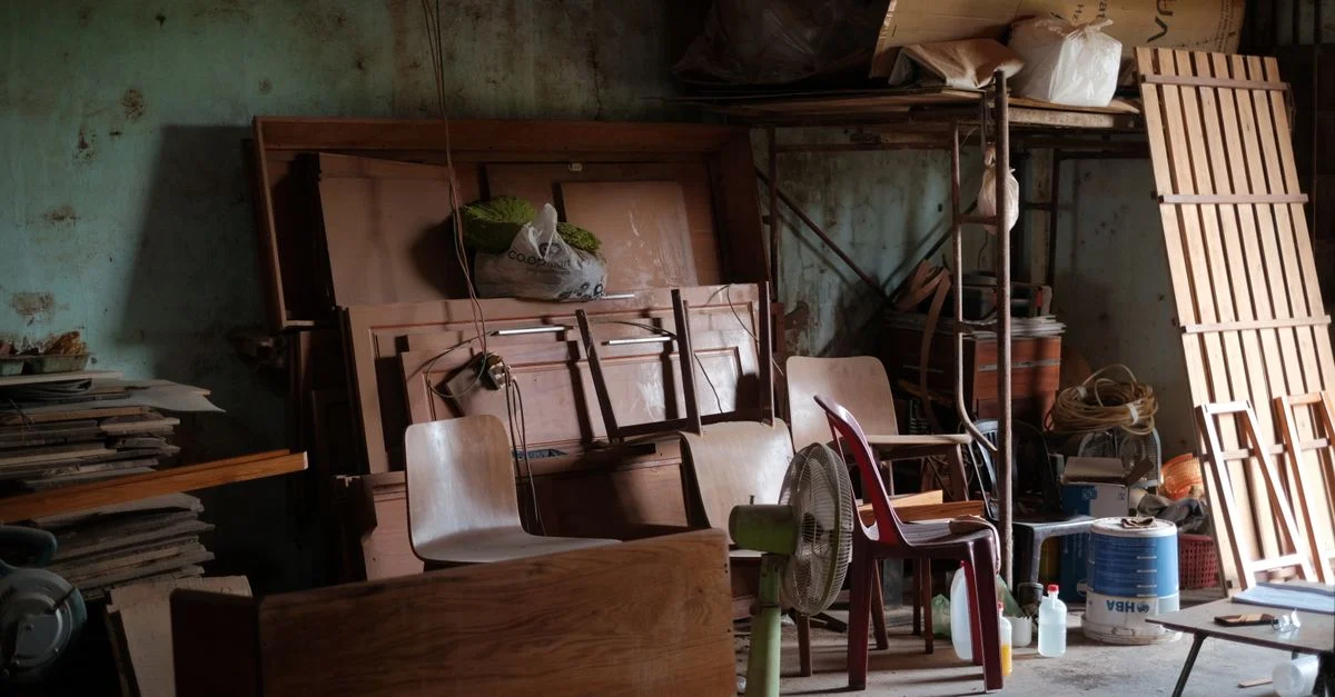 Un atelier encombré, rempli de meubles en bois, d'outils et d'objets divers, baigné de lumière naturelle.