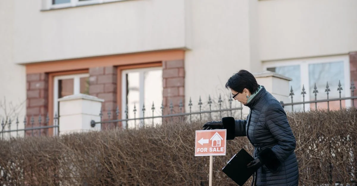 Agent immobilier en manteau noir plaçant un panneau 'À vendre' devant une maison.