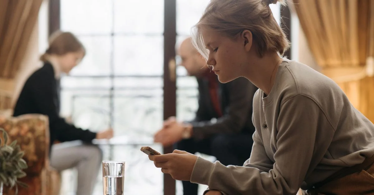 Adolescente concentrée sur son smartphone dans un cadre intérieur confortable avec des adultes flous en arrière-plan.