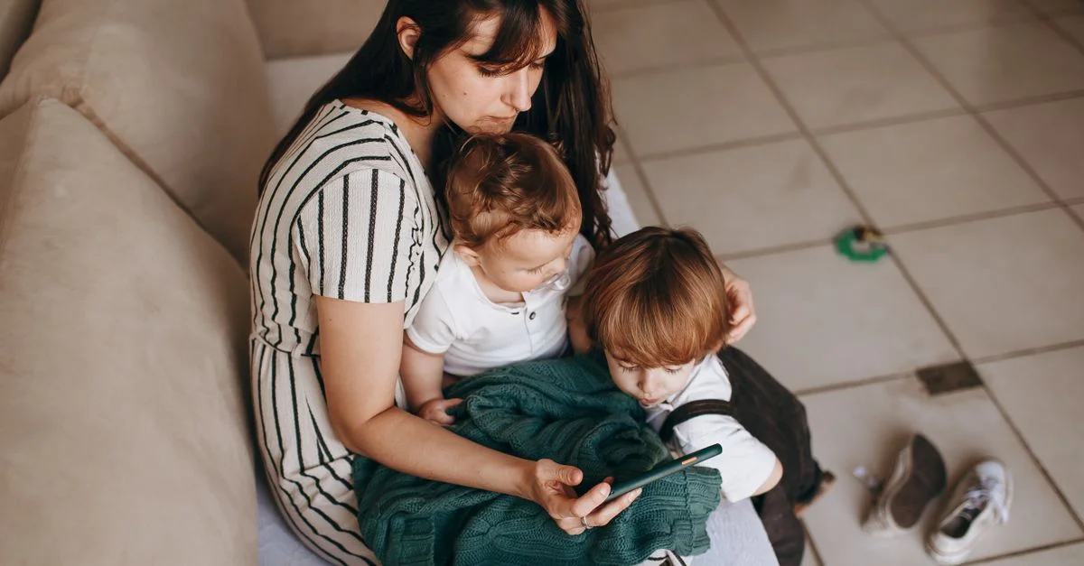 Une mère avec deux enfants assis sur le canapé, se rapprochant et utilisant un smartphone.