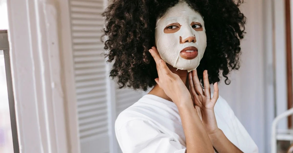 Une femme aux cheveux bouclés profite de soins de la peau avec un masque facial à l'intérieur, favorisant la détente et le bien-être.