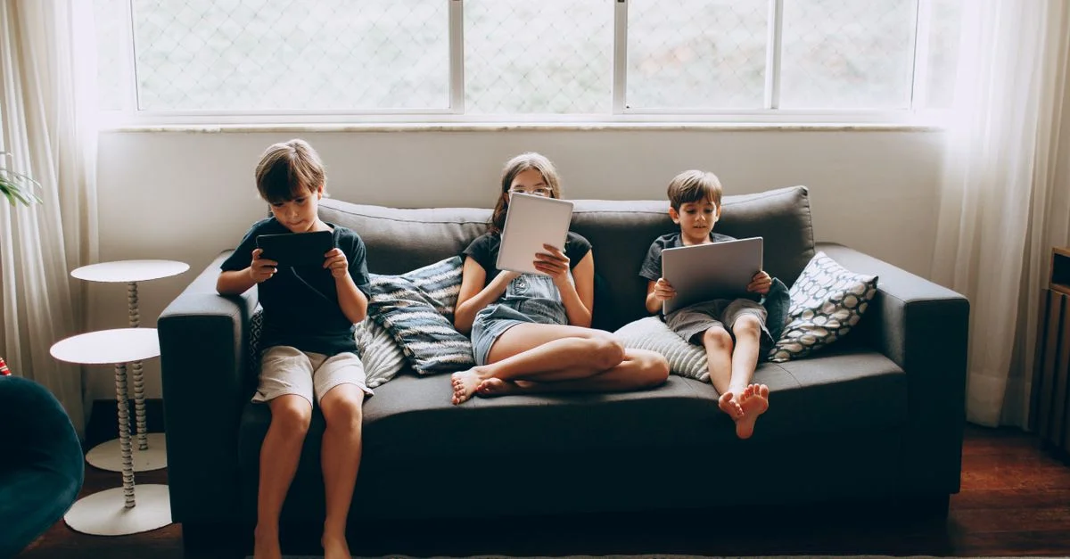Trois enfants se détendant sur un canapé avec des tablettes dans un salon lumineux et confortable.